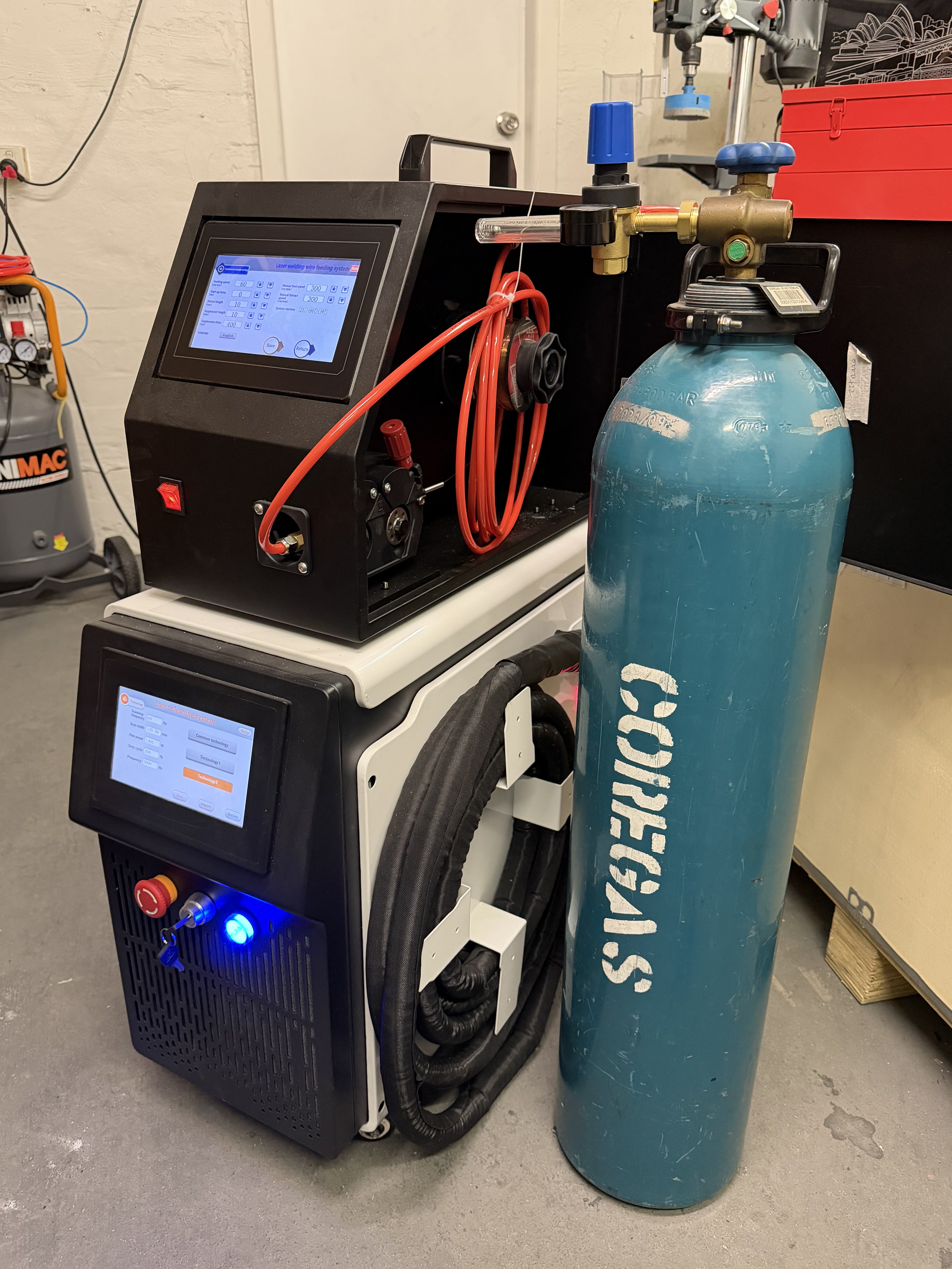 META MCW1500 handheld 3-in-1 laser system on the workshop floor — wire feeder unit on top with red welding cable, main 1500W air-cooled chassis below with screen lit and E-STOP, paired with a COREGAS argon shielding cylinder on the right